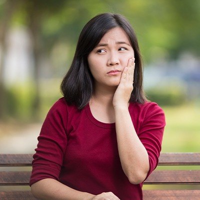 Woman sitting on bench holding cheek in pain before restorative dentistry