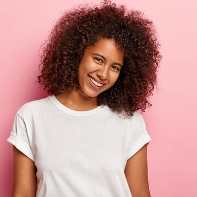 A young female wearing a white shirt and smiling against a pink background, showing off the small gap between her two front teeth in preparation to begin Invisalign treatment