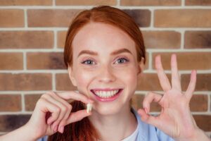 Woman smiling and holding extracted tooth while giving “OK” sign.